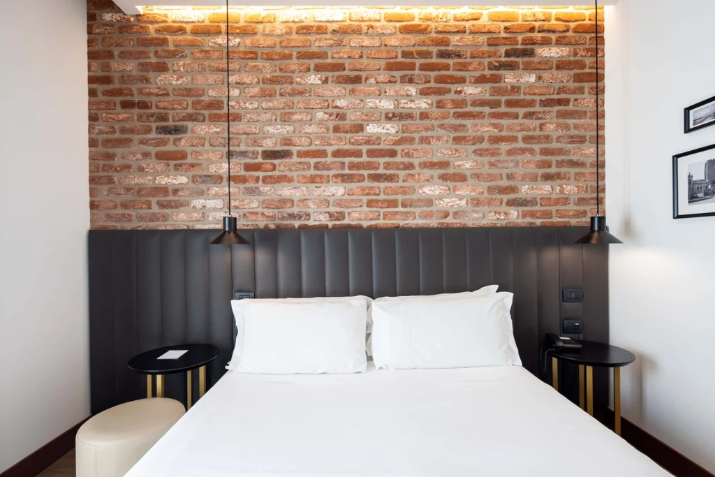 In the picture the superior room of the hotel with a double bed in the center and two small tables as bedside tables. The headboard of the bed is in black leather and the wall is made of bricks. From above hang two black pendant lamps.