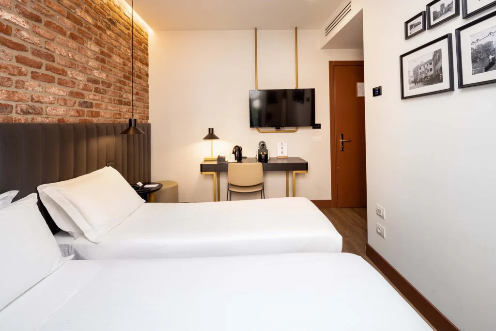 Nell'immagine la camera superior twin dell'hotel con due letti singoli, lunga testata nera in pelle e muro in mattoncini. Sulla parete in fondo c'è una scrivania nera con gambe oro con macchinetta caffè e bollitore e sedia. Sopra sul muro è appesa una tv e accanto c'è la porta d'ingresso della camera.
