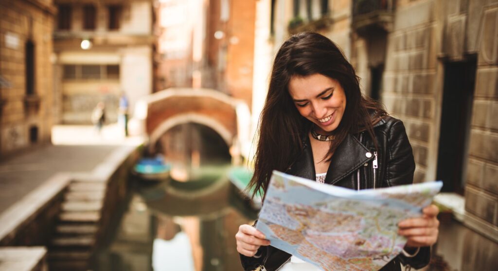 Nell'immagine si vede una ragazza che sorride e consulta una cartina. La ragazza si trova a Venezia, di fronte a un canale e uno dei tipici ponti della città.
