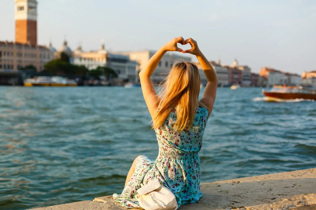 Nell'immagine una ragazza è seduta sul bordo di un canale e guarda di fronte a sè dove si vede una parte di Venezia. La ragazza ha le braccia sopra la testa unite per formare un cuore e indossa un abito azzurro con dei fiori rossi.
